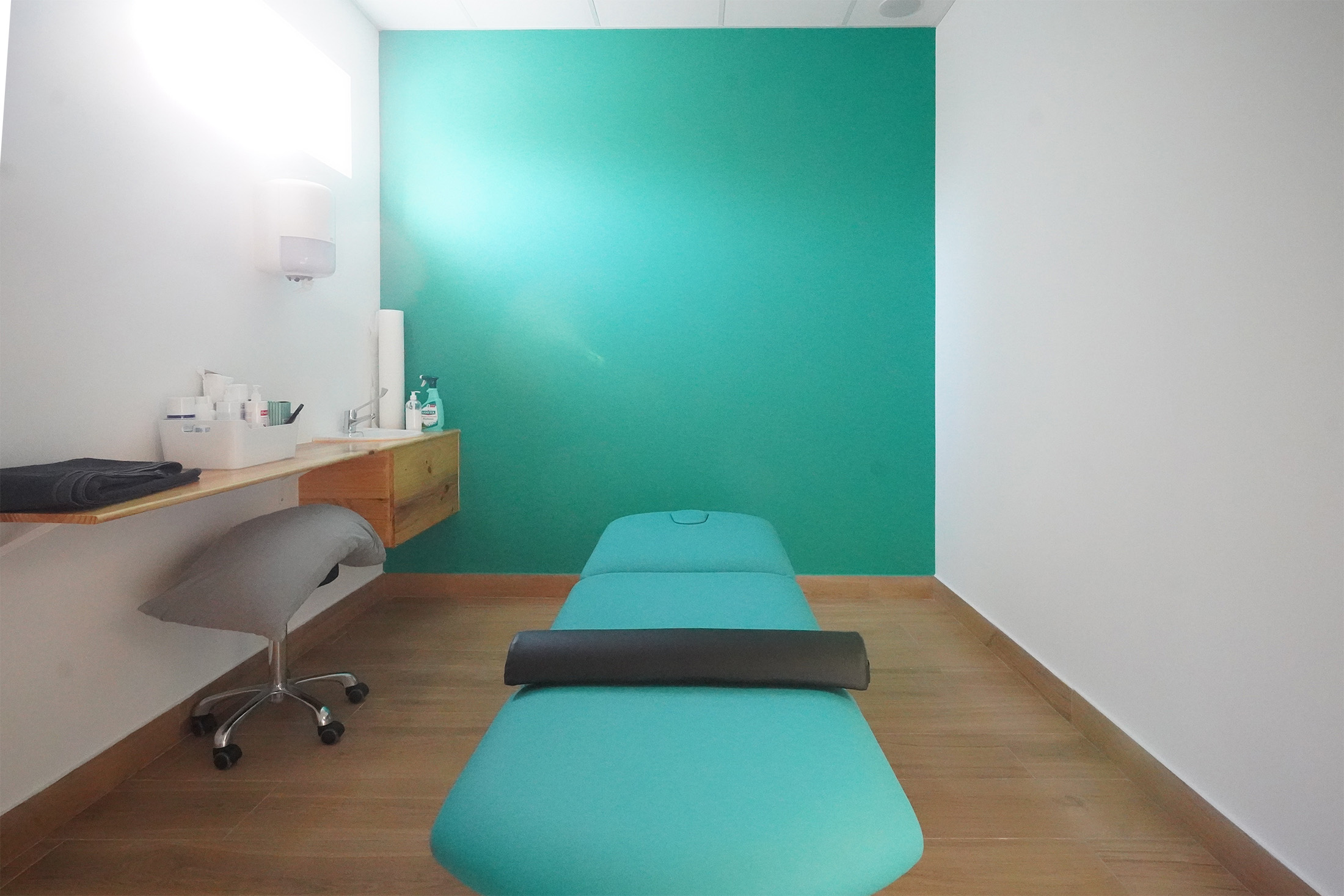 Sala de tratamiento o fisioterapia con camilla central, mueble de madera suspendido y pared turquesa